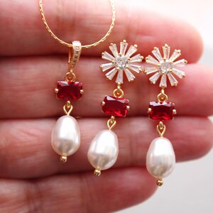 May include: A gold-toned jewelry set featuring a necklace and earrings. The necklace and earrings have red rectangular stones and white teardrop-shaped pearls. The earrings have a starburst design with clear crystals.