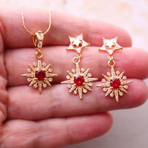 May include: Gold tone necklace and earrings set with a red gemstone starburst pendant and matching earrings. The earrings feature a smaller star charm above the starburst.