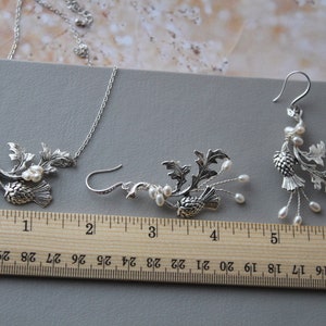 May include: A silver necklace and earrings set featuring a thistle design with pearl accents. The necklace is 18 inches long and the earrings are 2 inches long.