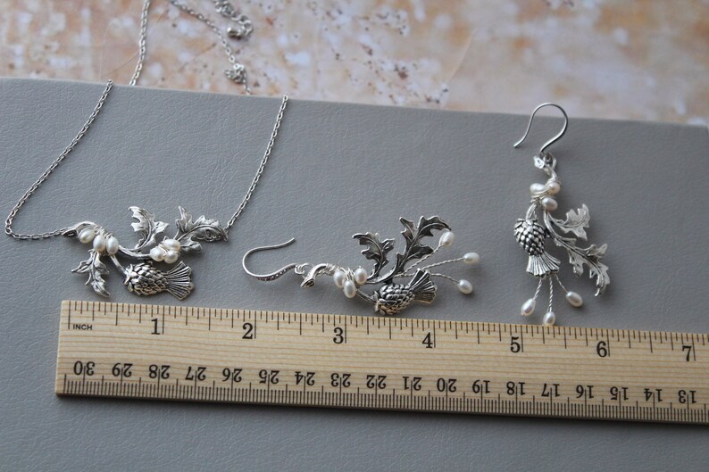May include: A silver necklace and earrings set featuring a thistle design with pearl accents. The necklace is 18 inches long and the earrings are 2 inches long.