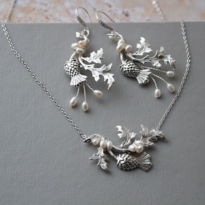 May include: A silver necklace and earrings set featuring a thistle design with pearl accents. The necklace has a delicate chain and a pendant with a thistle and leaves. The earrings are drop earrings with a thistle and leaves.