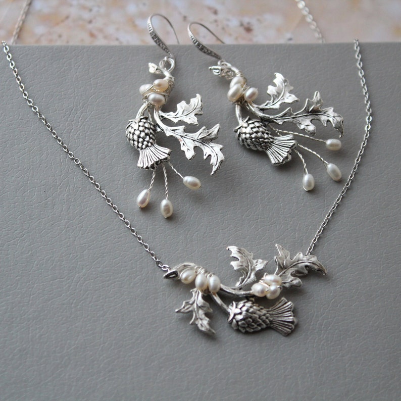 May include: A silver necklace and earrings set featuring a thistle design with pearl accents. The necklace has a delicate chain and a pendant with a thistle and leaves. The earrings are drop earrings with a thistle and leaves.