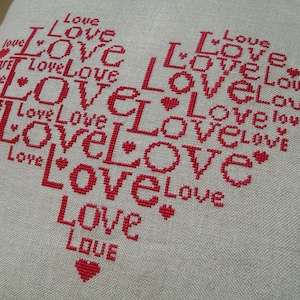 Pode incluir: Um desenho em forma de coração criado com fio de bordado vermelho sobre um fundo de tecido bege. O desenho é composto pela palavra "love" repetida várias vezes em tamanhos e orientações diferentes.