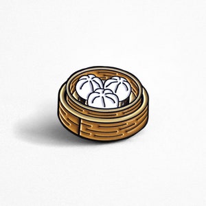 May include: A gold and brown enamel pin of a bamboo steamer with three white dumplings inside.