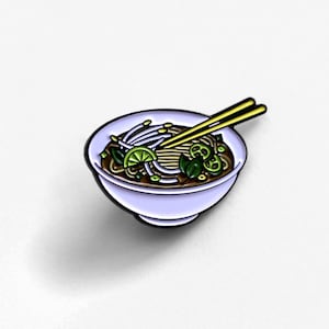 May include: A pin featuring a bowl of pho, a Vietnamese noodle soup. The pin has a light purple bowl with a black outline, filled with noodles, vegetables, and a lime wedge. Yellow chopsticks rest on top.