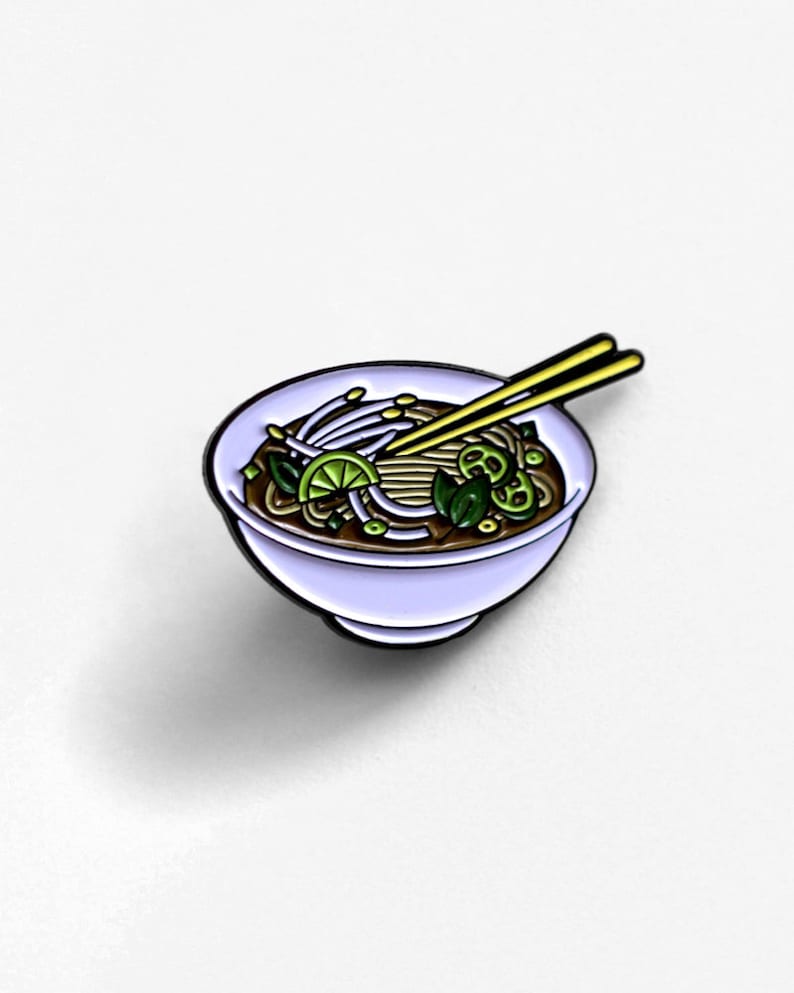 May include: A pin featuring a bowl of pho, a Vietnamese noodle soup. The pin has a light purple bowl with a black outline, filled with noodles, vegetables, and a lime wedge. Yellow chopsticks rest on top.