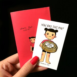 May include: A white card with the text "How Bao This Pin?" and a cartoon illustration of a person holding a basket of steamed buns. The card is on top of a red notebook with the text "What's that smell?" written on it.