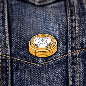 May include: A gold-colored metal pin with a cartoon image of a bamboo steamer filled with three white dumplings. The pin is on a blue denim background.