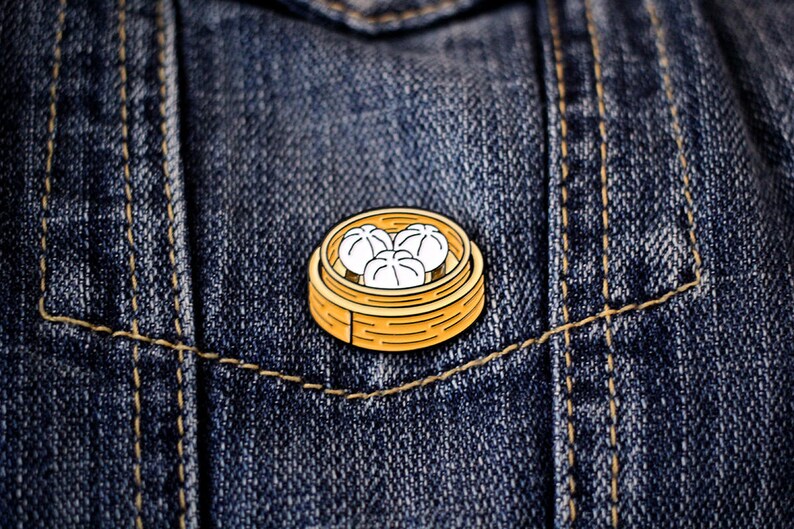 May include: A gold-colored metal pin with a cartoon image of a bamboo steamer filled with three white dumplings. The pin is on a blue denim background.