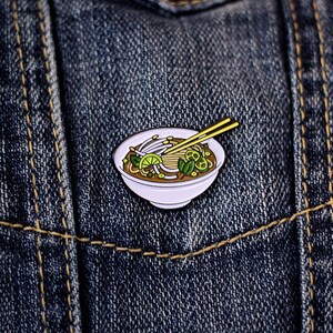 May include: Enamel pin depicting a bowl of pho with chopsticks. The pin features a white bowl filled with noodles, sprouts, lime, and herbs. The chopsticks are yellow. The pin is set against a blue denim background.