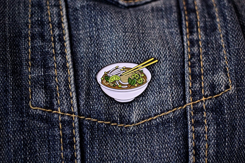 May include: Enamel pin depicting a bowl of pho with chopsticks. The pin features a white bowl filled with noodles, sprouts, lime, and herbs. The chopsticks are yellow. The pin is set against a blue denim background.