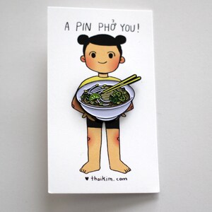 May include: A pin featuring a cartoon character holding a bowl of pho. The pin is on a white card with the text "A PIN PHỞ YOU!" above the character. The character is wearing a yellow shirt and black shorts. The website "thaikim.com" is at the bottom.