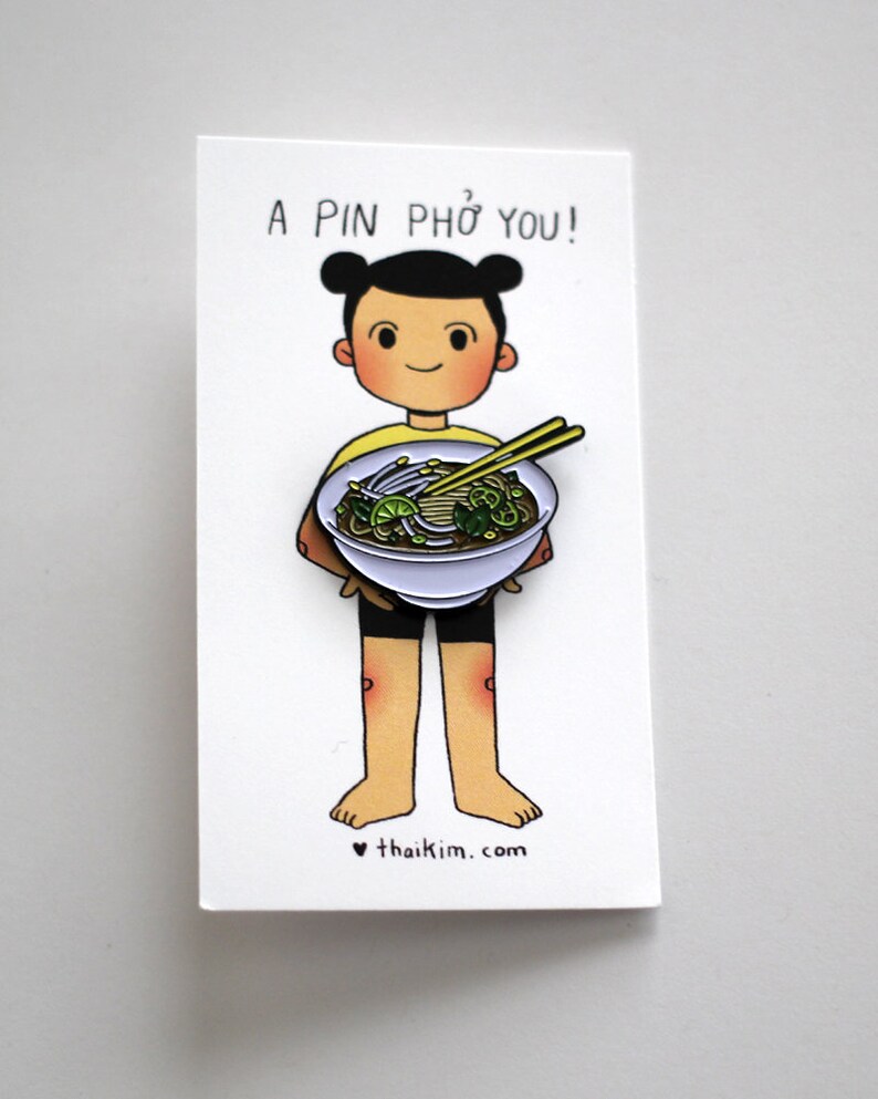May include: A pin featuring a cartoon character holding a bowl of pho. The pin is on a white card with the text "A PIN PHỞ YOU!" above the character. The character is wearing a yellow shirt and black shorts. The website "thaikim.com" is at the bottom.