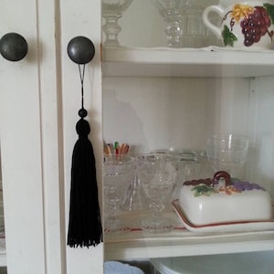 May include: A white cabinet with two dark grey knobs and a long black tassel hanging from one knob. Inside the cabinet are clear glasses, a white jug with a grape design, and a butter dish.