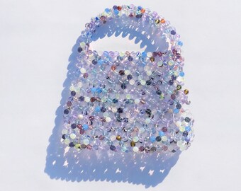May include: A small, beaded bag made of iridescent purple, blue, and white beads. The bag has a woven design and a top handle.