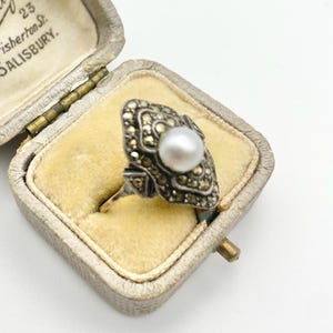 May include: Vintage silver ring with a large, round pearl center stone. The ring is set in a decorative, marcasite-studded setting. The ring is displayed in a vintage, cream-colored jewelry box with a yellow velvet interior.