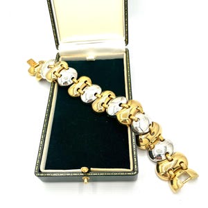 Puede incluir: Un brazalete dorado y plateado con un diseño de eslabones único, exhibido en una caja de joyería verde vintage. El brazalete presenta eslabones ovalados dorados y plateados alternados, creando un contraste llamativo. La caja tiene un borde dorado.