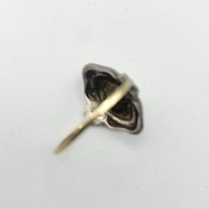 May include: A silver-toned ring with a gold band. The ring's face has a detailed, ornate design. The ring is set against a white background, showcasing its intricate details and craftsmanship. The ring's design is reminiscent of a flower.