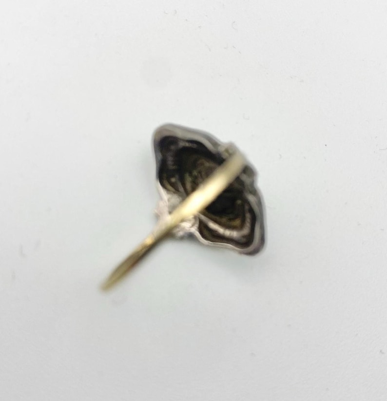 May include: A silver-toned ring with a gold band. The ring's face has a detailed, ornate design. The ring is set against a white background, showcasing its intricate details and craftsmanship. The ring's design is reminiscent of a flower.