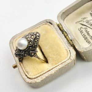 May include: An antique ring with a pearl and marcasite setting, displayed in an open, vintage jewelry box. The ring features a central pearl surrounded by ornate, dark-colored marcasite stones. The box is lined with a soft, yellow fabric.