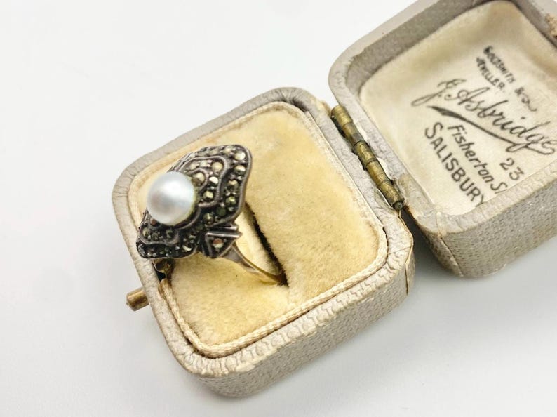 May include: An antique ring with a pearl and marcasite setting, displayed in an open, vintage jewelry box. The ring features a central pearl surrounded by ornate, dark-colored marcasite stones. The box is lined with a soft, yellow fabric.