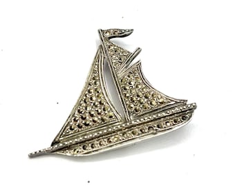May include: A silver-toned sailboat brooch featuring two sails and a hull decorated with numerous small, dark, faceted marcasite stones. The metalwork is polished, highlighting the detailed structure of the boat, including a small flag at the top.