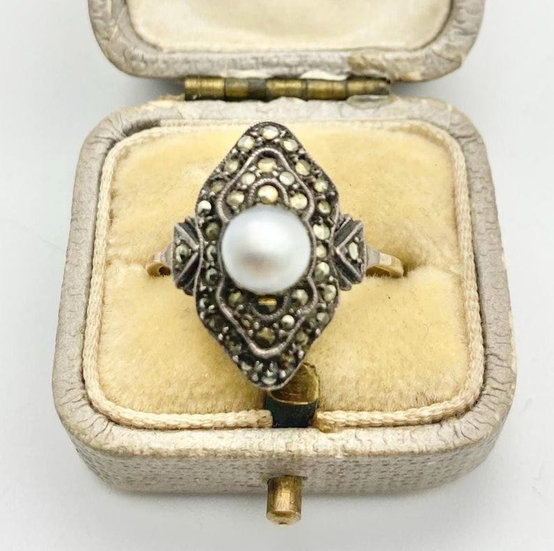 May include: Vintage ring with a central pearl, surrounded by marcasite stones in a decorative setting. The ring is set in a gold band and displayed in a vintage jewelry box with a yellow velvet interior. A classic piece of jewelry.