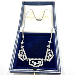 Puede incluir: Un collar de plata con diseño geométrico, engastado con piedras preciosas transparentes, presentado en una caja azul con la marca "Mappin & Webb". El collar presenta formas florales y geométricas.