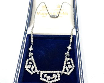 May include: A silver necklace with a geometric design, set with clear gemstones, displayed in a blue box with the brand name "Mappin & Webb". The necklace features floral and geometric shapes.