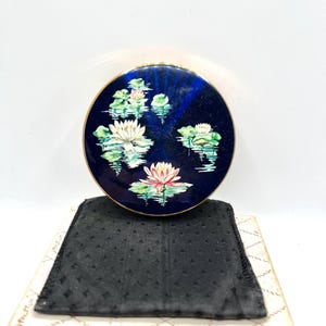 May include: A round, navy blue compact with a gold rim, featuring a painted water lily design. The design includes white and pink flowers with green lily pads on a reflective water surface. Includes a black pouch and a patterned box.