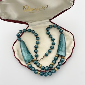 May include: A teal blue beaded necklace with two large teal blue pendant charms in a red and white shell-shaped box. The box has the text "Rosita Pearl Charm" on the inside lid.