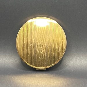 May include: A round, gold-colored compact with a textured, striped design.