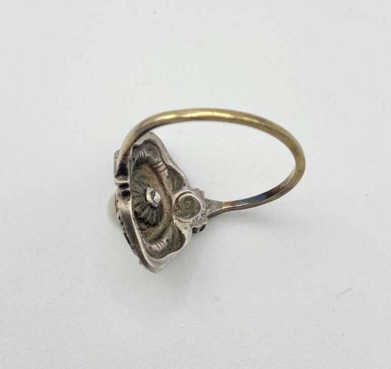 May include: Vintage silver ring with a gold band. The ring features an ornate, detailed setting with a central pearl and small accent stones. The setting has an intricate floral design.