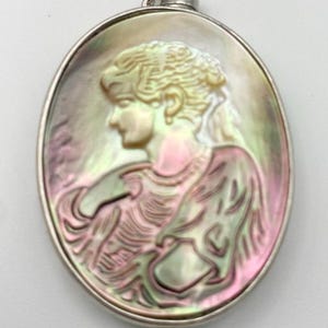 May include: An oval-shaped pendant with a silver-toned frame and a carved mother-of-pearl design of a woman's profile. The woman's hair is styled in an updo and she is wearing a necklace.