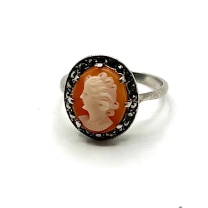 Può includere: Anello con cammeo ovale raffigurante il profilo di una donna in color pesca chiaro. Il cammeo è incastonato in un anello in metallo argentato con un bordo di pietre nere. Un gioiello classico.