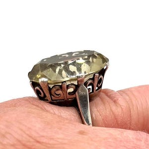 May include: A ring featuring a large, oval-cut, pale yellow gemstone set in a silver-toned metal basket setting. The setting displays an intricate, openwork scroll pattern along the sides, supporting the faceted stone securely in place.
