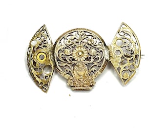 May include: A gold-toned metal brooch featuring an intricate filigree design. The central circular element displays a floral motif above a vase shape, flanked by two symmetrical, fan-shaped side panels with openwork scroll and shell patterns.