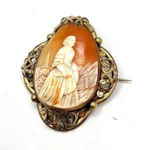 May include: Antique cameo brooch depicting a carved woman in a white dress against a landscape. The oval cameo is set in a gold-toned filigree frame with a pin. The cameo has a warm, orange-brown colour.