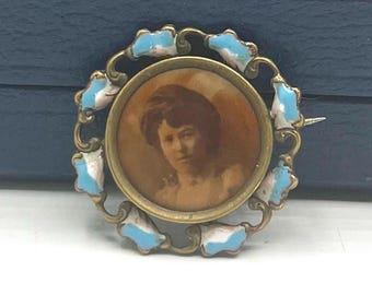 May include: Antique gold brooch featuring a portrait of a woman. The circular frame is adorned with light blue and white enamel details. A vintage jewellery item.