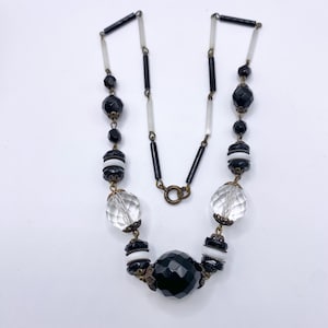 Puede incluir: Un collar de cuentas negras y blancas con una gran cuenta negra facetada en el centro. El collar está hecho con cuentas negras y blancas alternas, y tiene una cadena simple.