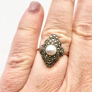 May include: A vintage gold-tone ring featuring a central white pearl. The ring's setting is an ornate, elongated shape, encrusted with small, sparkling marcasite stones. The ring is on a finger.