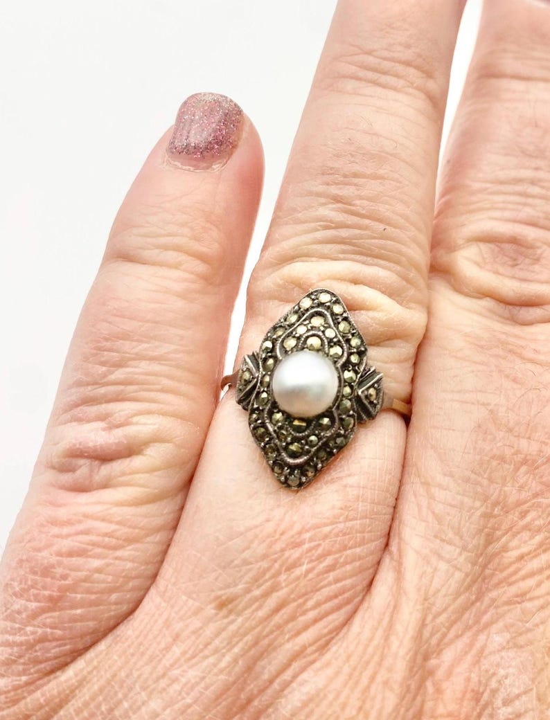May include: A vintage gold-tone ring featuring a central white pearl. The ring's setting is an ornate, elongated shape, encrusted with small, sparkling marcasite stones. The ring is on a finger.