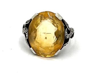 May include: A ring featuring a large, oval-cut, translucent yellow gemstone held by four silver prongs. The band is crafted from dark, oxidized silver with intricate, swirling filigree patterns along the sides, providing a contrast to the stone.