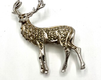 May include: A silver-toned deer brooch with detailed antlers and a textured body. The brooch is set against a white background, showcasing its intricate design and craftsmanship. The deer is standing, with its legs extended.