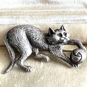 Könnte beinhalten: Eine silberfarbene Metallbrosche in Form einer springenden Katze. Die Katze hat eine detaillierte Fellstruktur und streckt ihre Vorderpfoten aus, um ein kleines, rundes Wollknäuel zu berühren. Die Brosche ist auf der Rückseite mit einer Nadel versehen.