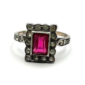 Peut inclure: Une bague argentée ornée d'une pierre centrale rectangulaire rose vif taille émeraude. La pierre centrale est entourée d'une bordure festonnée de petites pierres claires taille ronde. L'anneau métallique présente aussi des pierres claires.