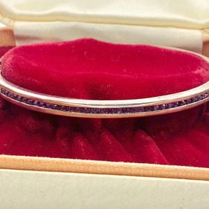 May include: A silver-coloured hinged bangle bracelet with a row of small, dark purple gemstones. The bracelet is displayed inside a vintage, cream-coloured jewellery box with a red velvet interior.