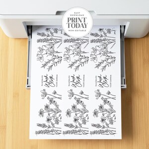 Op de afbeelding: Een wit vel papier dat uit een printer komt, met drie identieke verticale labels met zwarte lijntekeningen van wilde bloemen. Elk label bevat de tekst "Handmade Wash Cloth 100% Cotton" in een combinatie van script en schreefloos lettertype.