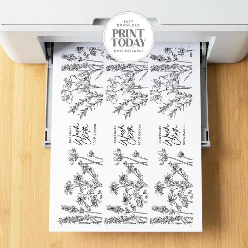 Op de afbeelding: Een wit vel papier dat uit een printer komt, met drie identieke verticale labels met zwarte lijntekeningen van wilde bloemen. Elk label bevat de tekst "Handmade Wash Cloth 100% Cotton" in een combinatie van script en schreefloos lettertype.