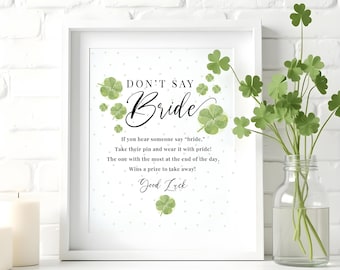 May include: A framed print with the text "DON'T SAY Bride" and instructions for a bridal shower game. The print is decorated with green shamrocks and a white background. A vase of shamrocks is to the right.
